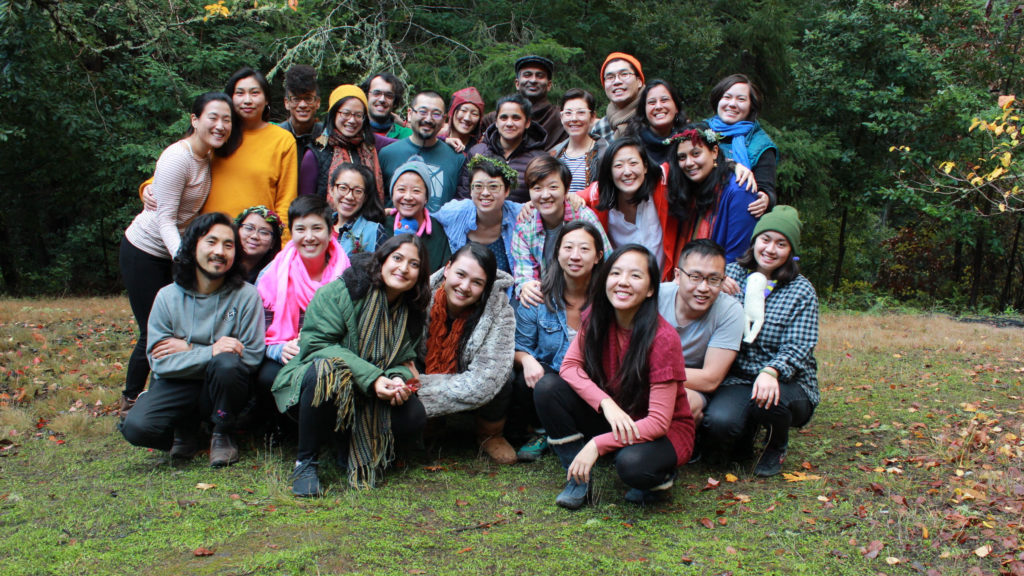 Image description: 28 participants of the 2018 API Jam gather outside on the grass. There are 3 row of people some standing, some kneeling, all smiling, and many with their arms around each other.